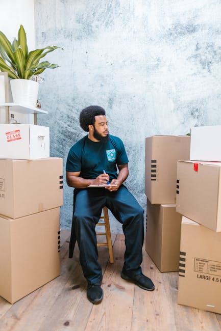 A young male mover dressed in a navy blue uniform and black bandana with white patterns, standing inside a property amidst packed cardboard boxes and packing materials. He is holding a clipboard and p